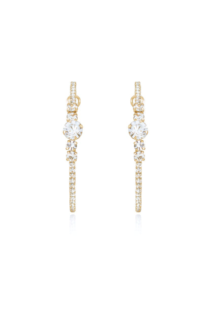 Gold earrings adorned with sparkling crystals hang vertically, catching light and reflecting it. They are displayed against a white background, emphasizing their elegance and shine.