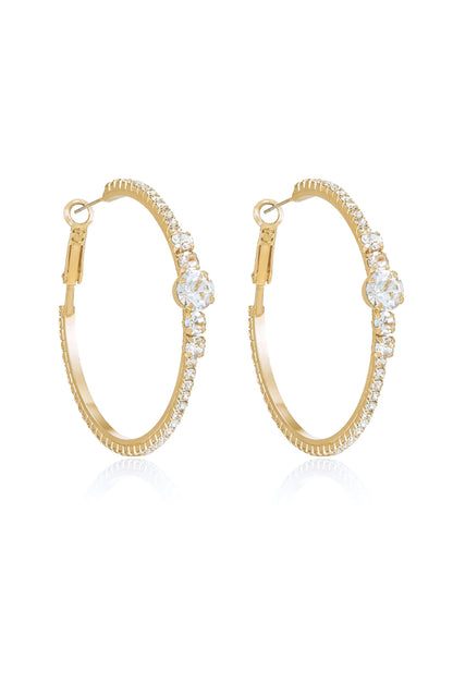 A pair of gold hoop earrings with sparkling clear stones along the outer edge and a larger stone prominently featured at the center, set against a reflective white background.