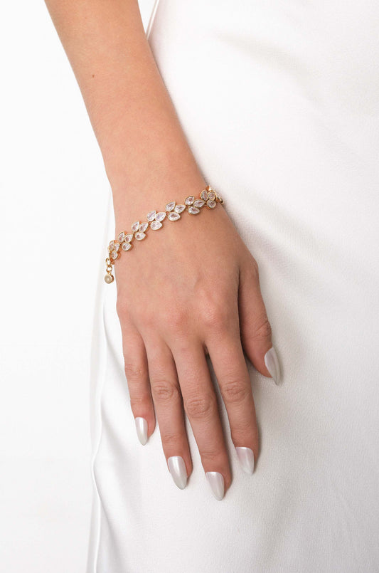 A hand adorned with a gold bracelet featuring leaf-shaped, clear gemstones rests on a silky, white fabric surface, highlighting the elegance of both the jewelry and the setting.