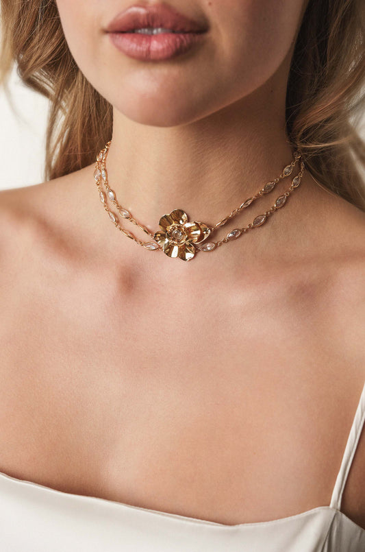 A golden flower necklace is elegantly displayed around a woman's neck, accentuating her collarbone, while she looks toward the camera with slightly parted lips, set against a soft, neutral background.