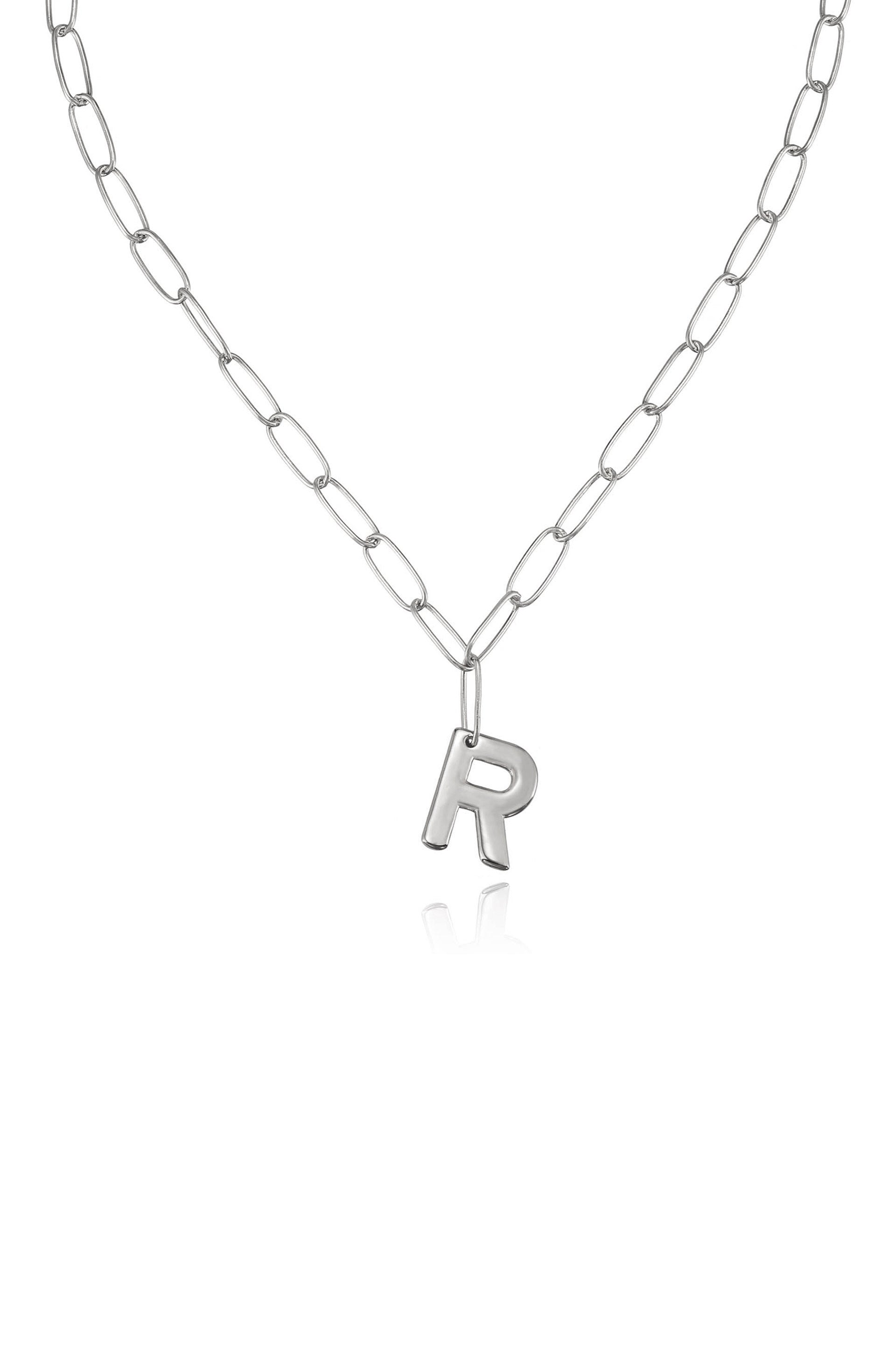 A silver chain necklace features large oval links, with a shiny letter "R" charm hanging from it. The necklace rests against a neutral background, reflecting light subtly.