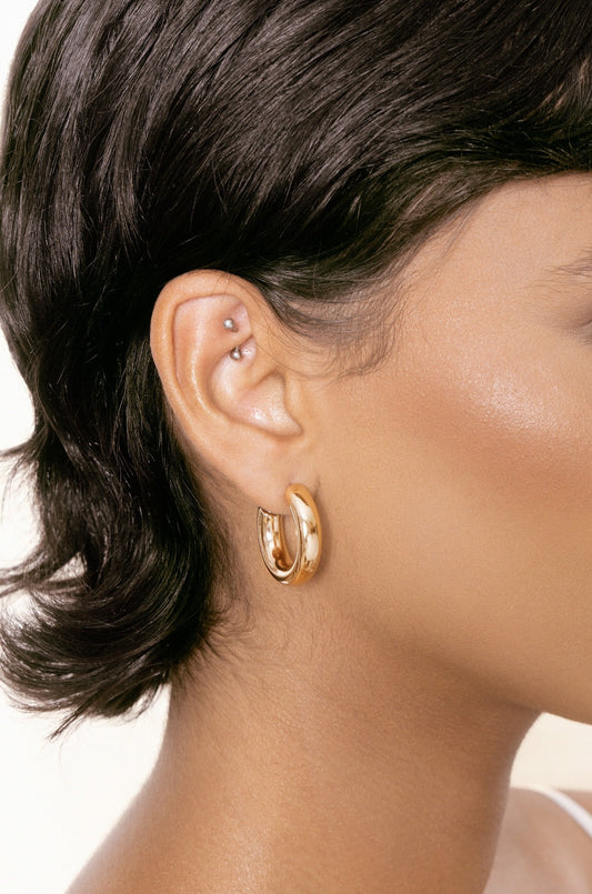 A close-up of a person's ear adorned with a shiny gold hoop earring. The smooth skin contrasts with the dark, wavy hair, and a small piercing is visible above the earring.