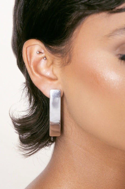 A close-up of a person's ear featuring a large, shiny silver earring. The individual has short, dark hair and subtle makeup, set against a soft, light-colored background.