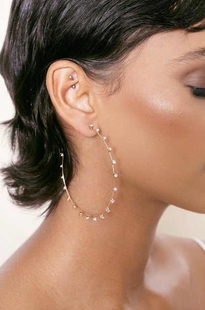 A large, delicate gold hoop earring, adorned with small gemstones, hangs from the lobe of a close-cropped hairstyle, accentuating the smooth skin and ear piercings against a soft background.