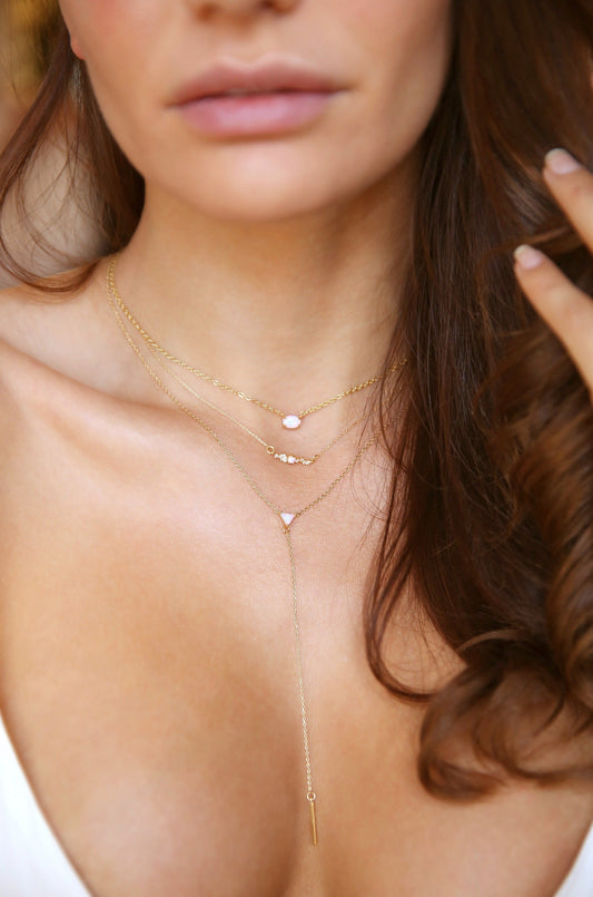 Elegant layered gold necklaces adorned with small gemstones rest on the neck and collarbone of a person, who gently touches their hair, set against a soft, blurred background.