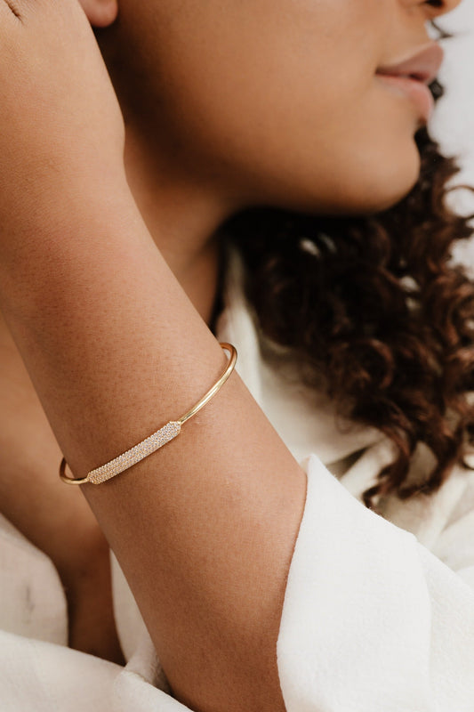 A gold bracelet adorned with a sparkling detail rests on a person's tanned wrist. The individual is partially visible, wearing a light-colored garment and has curly hair.