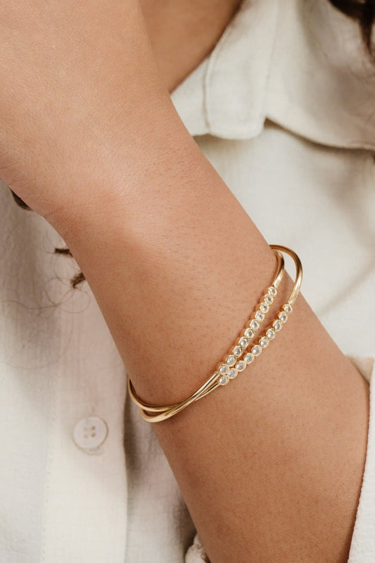 A gold bracelet with clear gemstones is resting on a person's wrist. The person is wearing a light-colored shirt, and their skin tone is visible in the close-up.