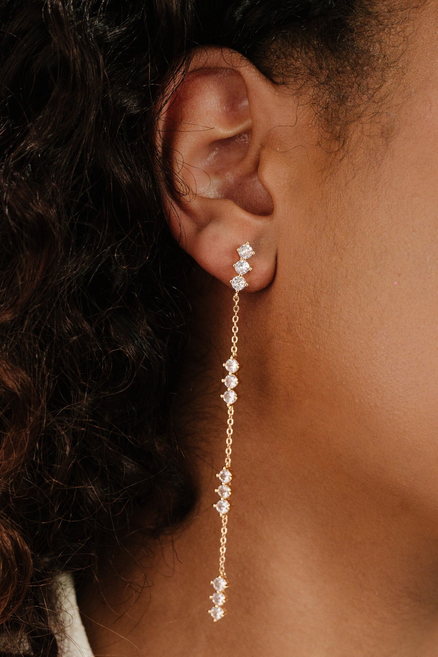 A gold dangle earring adorned with multiple sparkling gemstones hangs from the earlobe of a person with curly hair. The skin tone appears warm and the setting is neutral.