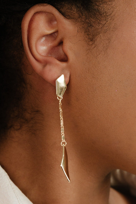 A gold earring hangs delicately from a person's ear, featuring a geometric design and a chain. The earring's shiny surface contrasts against the skin tone in a softly lit setting.