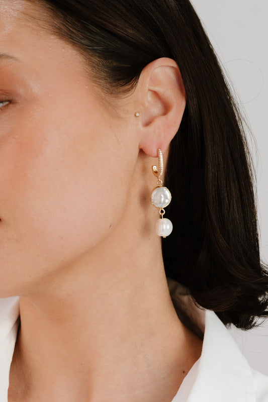 A close-up of a woman's ear showcases a gold hoop earring adorned with two pearls, one larger and round, the other smaller, set against a neutral background.