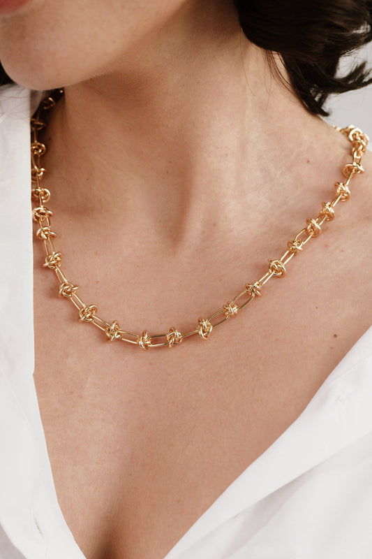 A gold necklace adorns a woman's neck, featuring interlinked circular designs. The individual is partially visible, dressed in a white shirt, with a soft, neutral background.