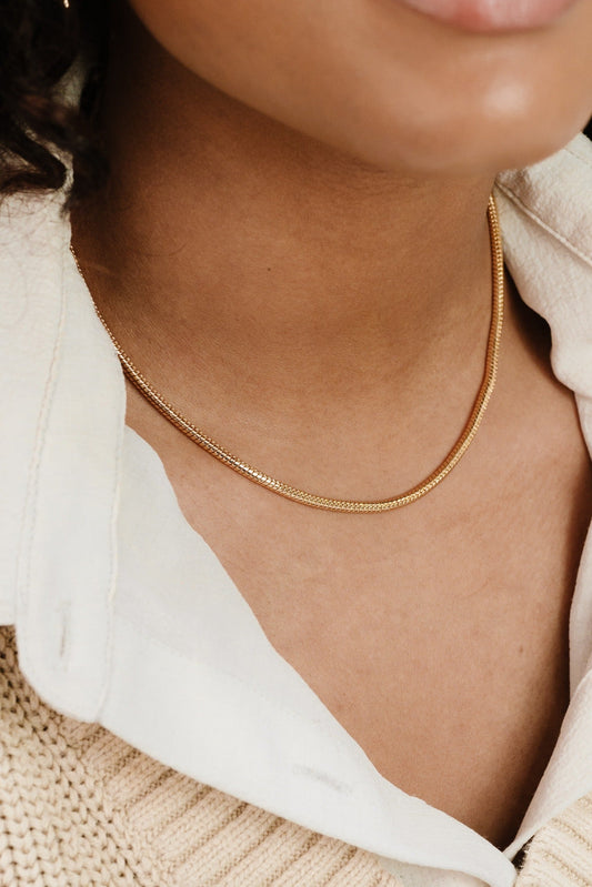 A gold necklace rests gently on a person's neck, accentuating their skin tone. The individual wears a white shirt collar, partially visible against a knitted beige garment.