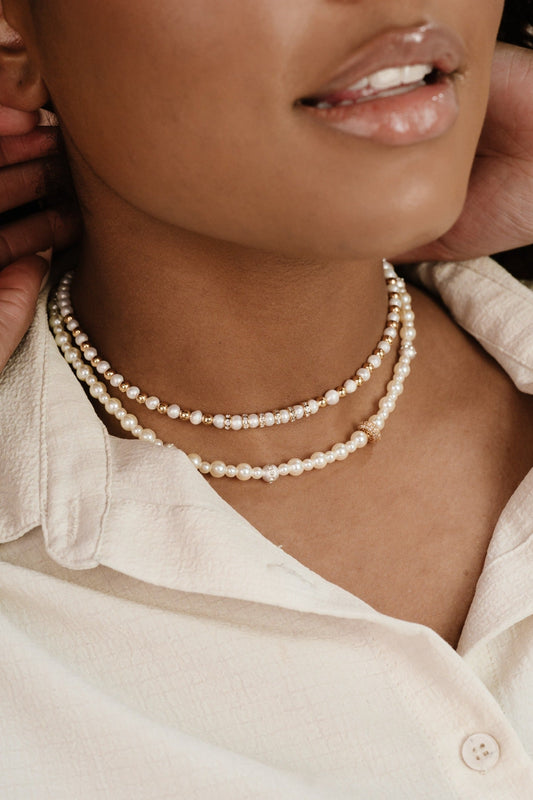 A close-up of a person's neck features multiple layers of pearl necklaces. A hand adjusts one necklace while the person, wearing a light shirt, smiles gently. The setting appears casual and intimate.