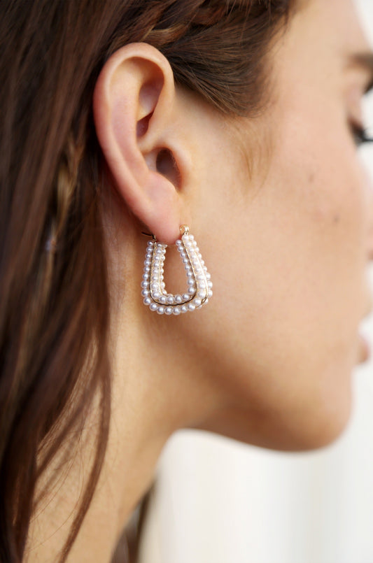 A large, decorative earring hangs from a womanâ€™s ear, featuring a double-layered design with small pearls. The background is softly blurred, focusing on the earring and the profile of her face.