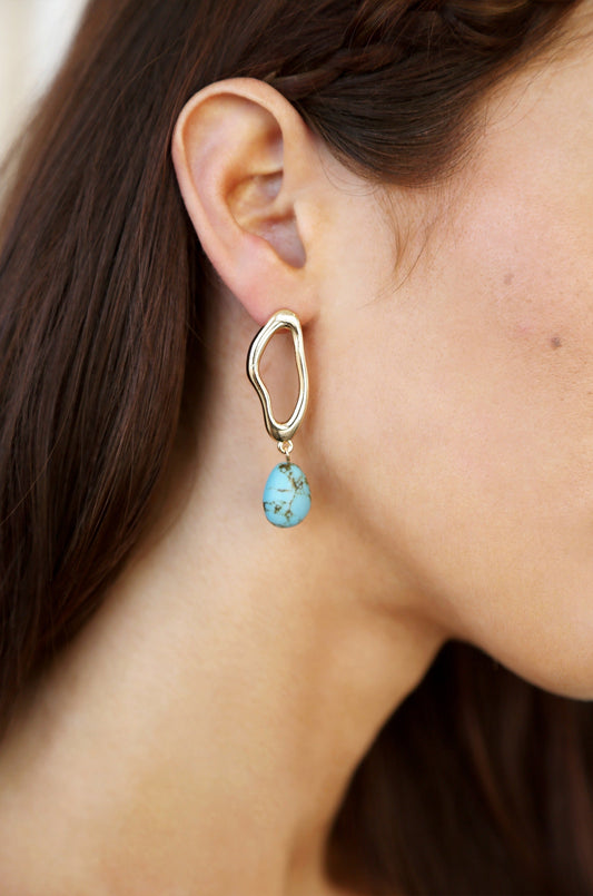 A golden earring with an abstract shape showcases a turquoise stone hanging below it. The earring is worn on a woman's ear, highlighting her smooth skin and brown hair.