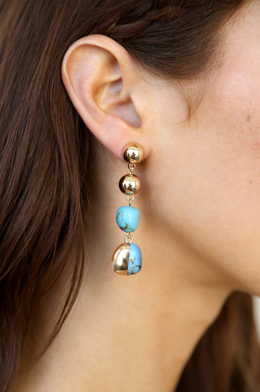 Gold and turquoise earrings dangle from a woman's ear, with three elements: two shiny gold spheres above and a combination of blue stones and gold below, enhancing her profile.