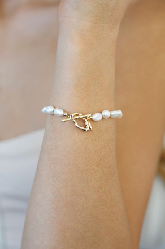 A close-up of a wrist adorned with a delicate bracelet featuring white pearls and a twisted gold clasp. The skin tone suggests a soft and natural ambiance, enhancing the jewelry's elegance.