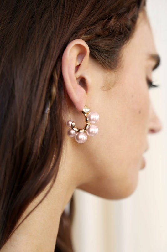 A close-up of a woman's ear adorned with a stylish earring featuring pink pearls and a gold accent, set against a softly blurred, neutral background.