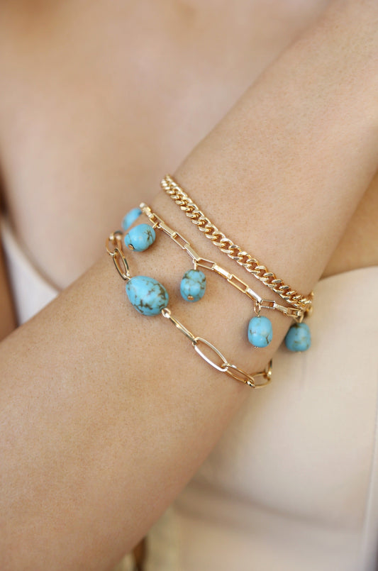 A close-up of a womanâ€™s arm adorned with three bracelets; one gold chain and two featuring turquoise beads, highlighting a stylish accessory choice against a neutral background.