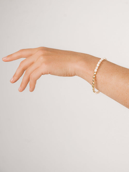 A hand, with fingers slightly curled and extending outward, displays a bracelet made of alternating white beads and gold accents. The background is a soft, light gradient.