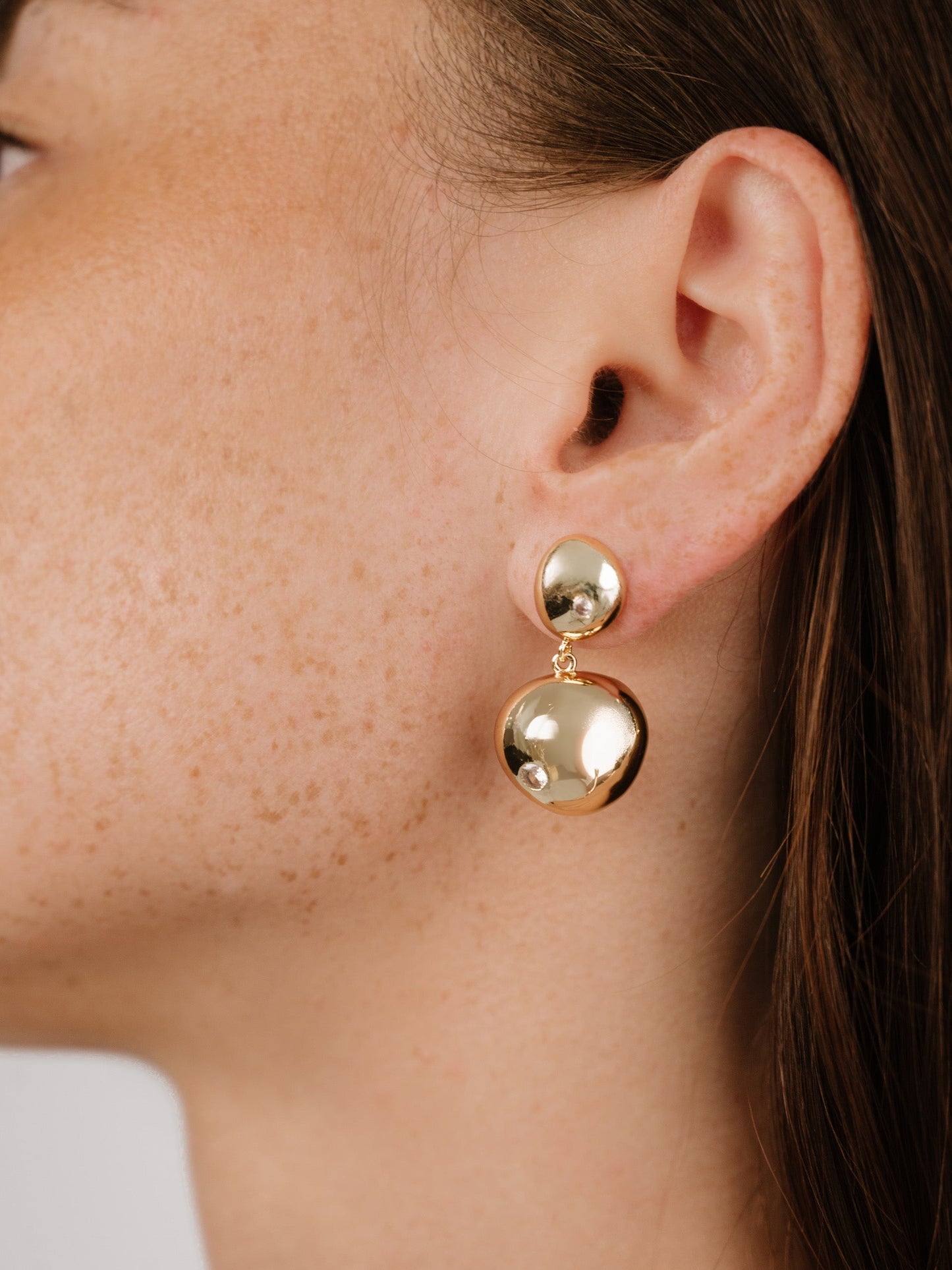 A person's ear showcases a pair of gold earringsâ€”one round and one elongatedâ€”reflecting light. The focus is on the jewelry against their lightly freckled skin and brown hair.