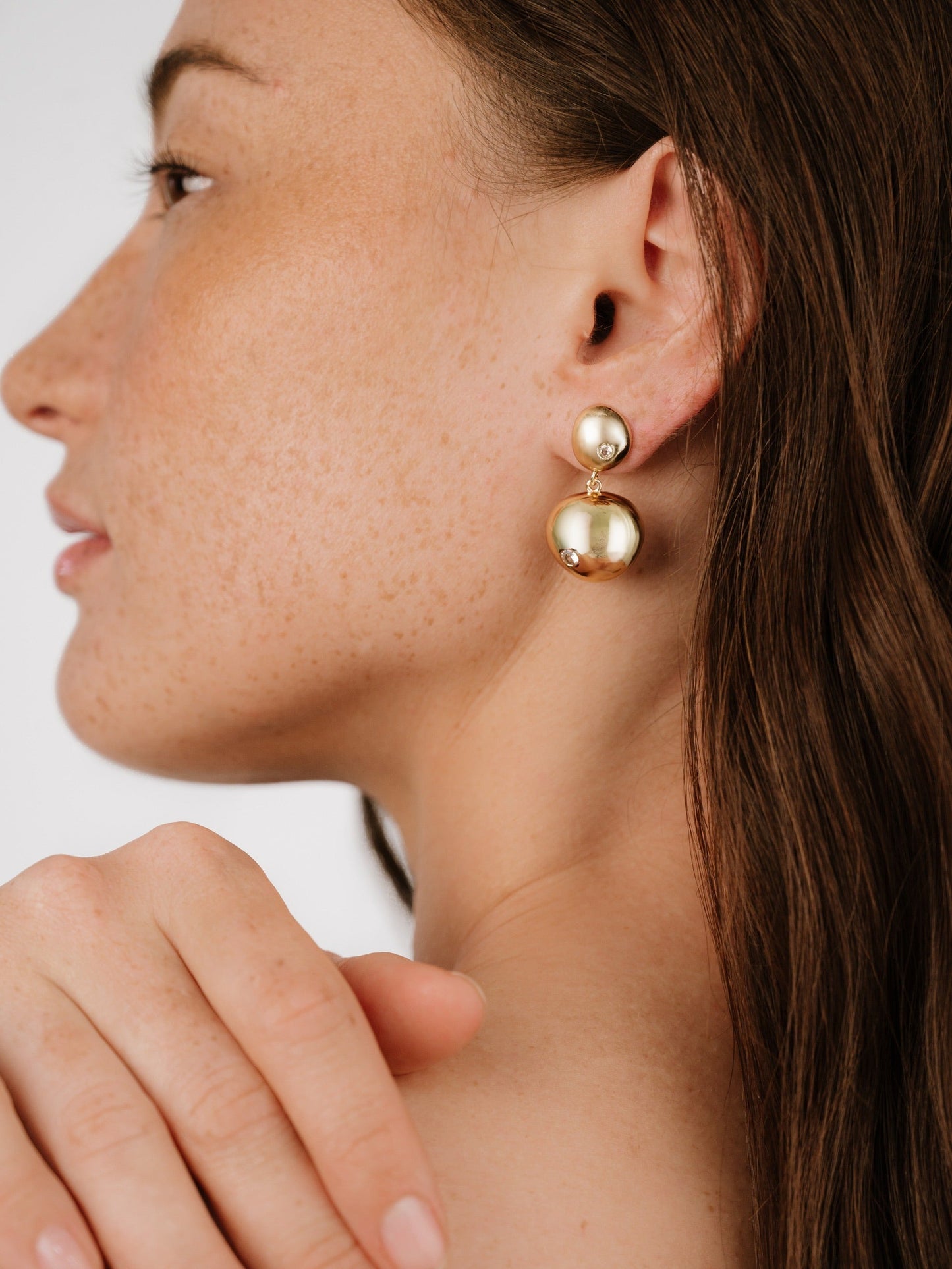 A person with a light complexion and freckles wears large gold earrings. She gently touches her shoulder, with soft lighting highlighting her profile against a plain background.