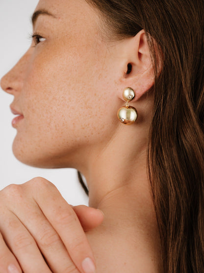 A person with a light complexion and freckles wears large gold earrings. She gently touches her shoulder, with soft lighting highlighting her profile against a plain background.