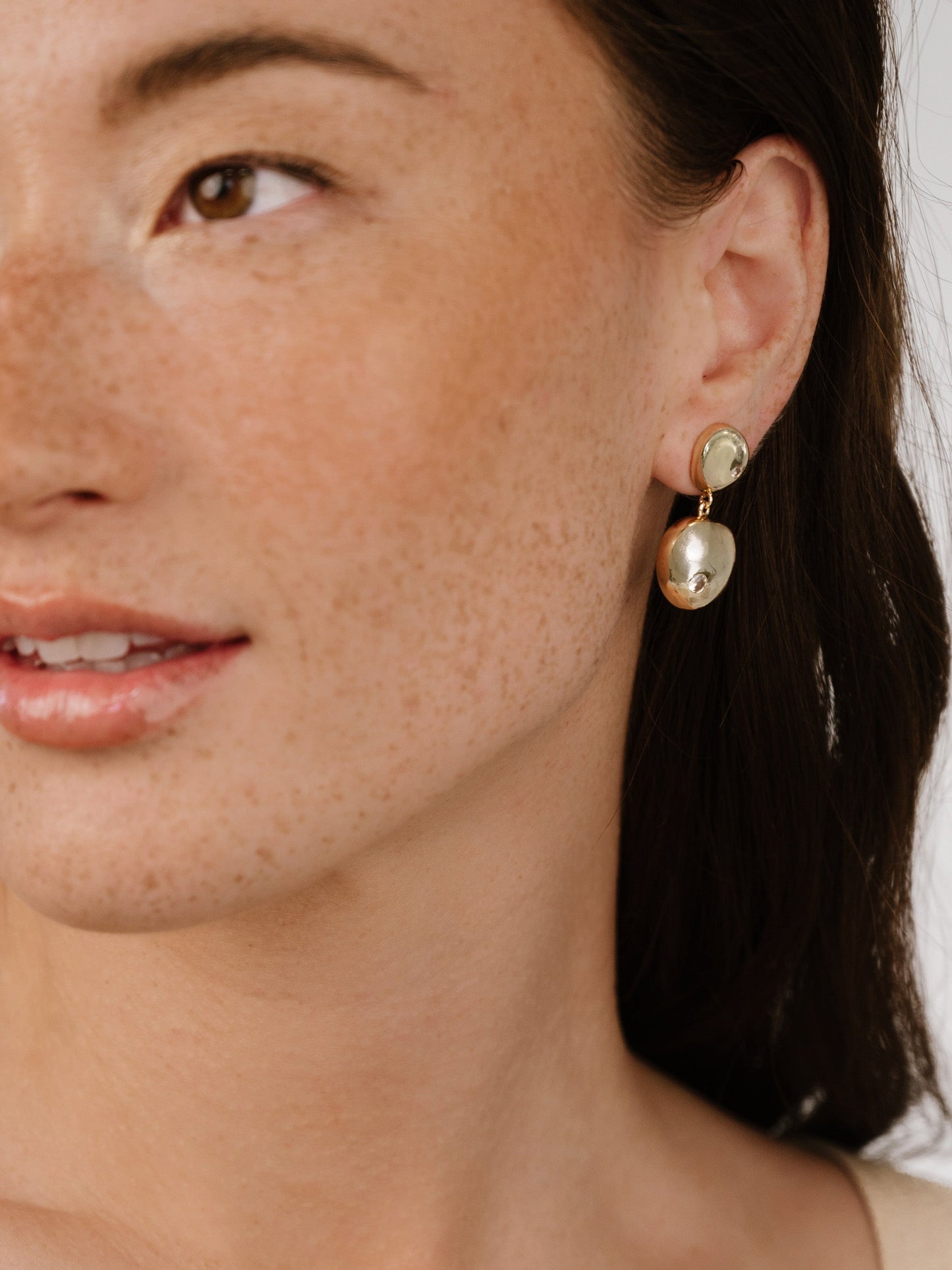 A close-up of a womanâ€™s face showcases her wearing a pair of elegant earrings. She smiles softly, surrounded by a neutral background that highlights her features and accessories.