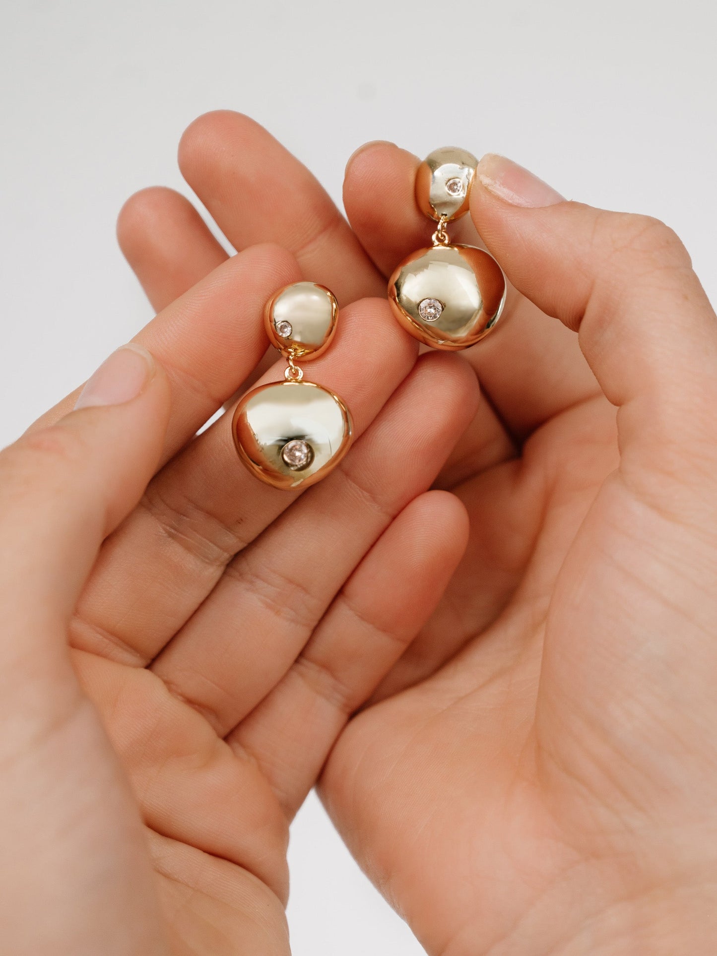 A pair of gold-tone earrings, featuring rounded shapes and sparkling stones, is being held between two hands against a light-colored background, showcasing their elegant design and shine.