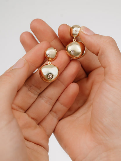 A pair of gold-tone earrings, featuring rounded shapes and sparkling stones, is being held between two hands against a light-colored background, showcasing their elegant design and shine.