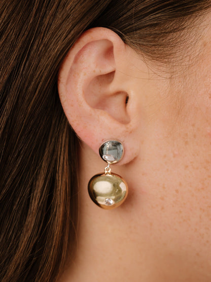 A pair of stylish earrings is showcased, featuring a dark, glossy stone at the top and a shiny, gold-colored orb below, hanging elegantly from the lobe of a human ear.