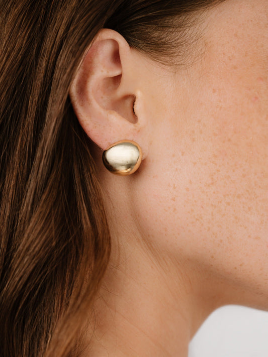 A gold-colored earrings is worn on the ear of a person with light brown hair, showcasing the earringâ€™s polished surface against a background of soft, neutral tones.