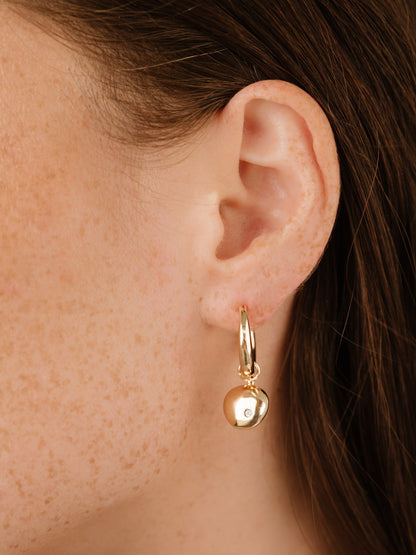 A gold hoop earring with a dangling, round charm is prominently displayed near a woman's ear, showcasing her freckled skin and dark hair in a soft lighting context.