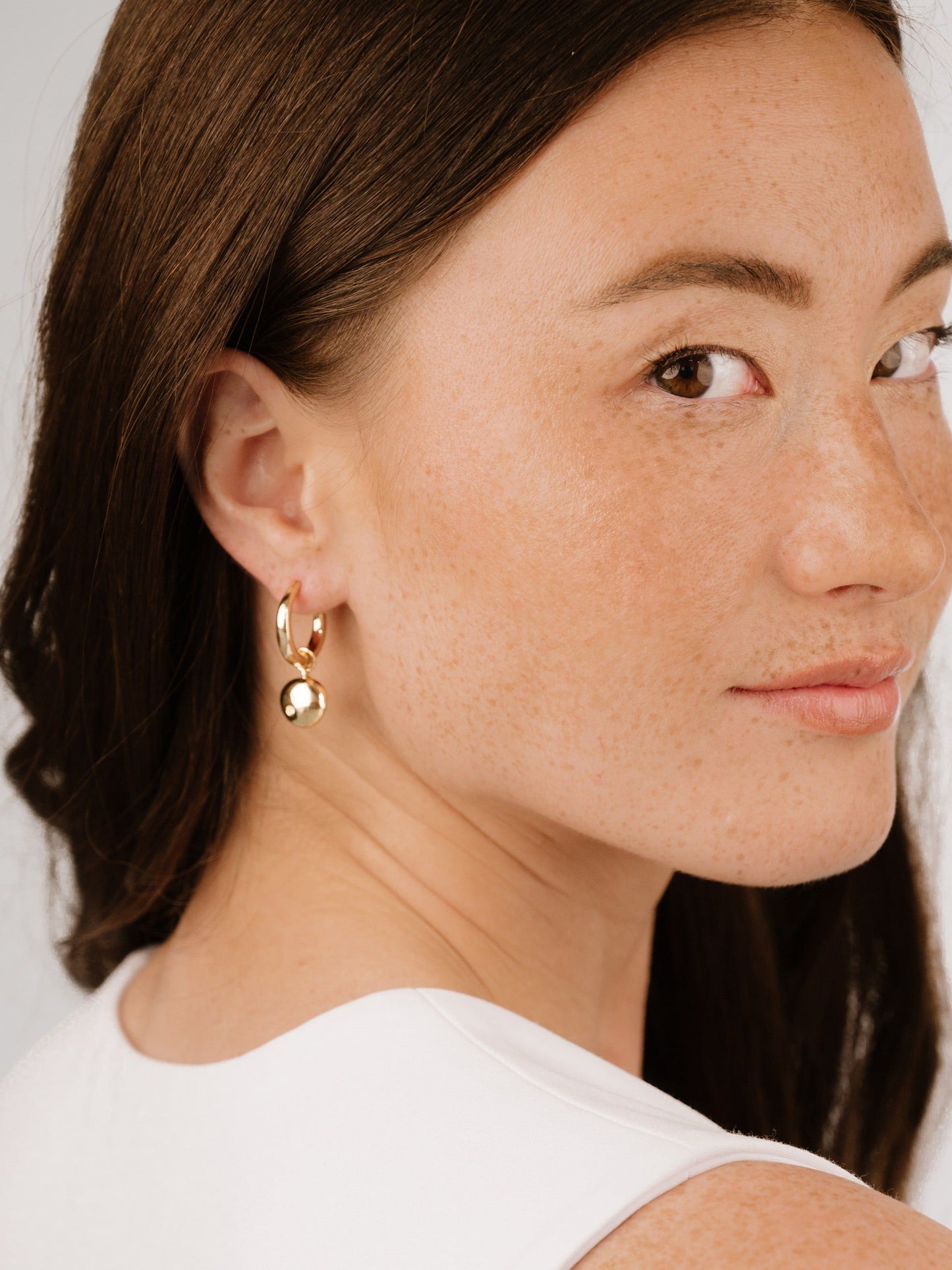 A woman with smooth, brown hair is wearing a gold hoop earring featuring a round charm, looking over her shoulder with a soft expression against a light, neutral background.