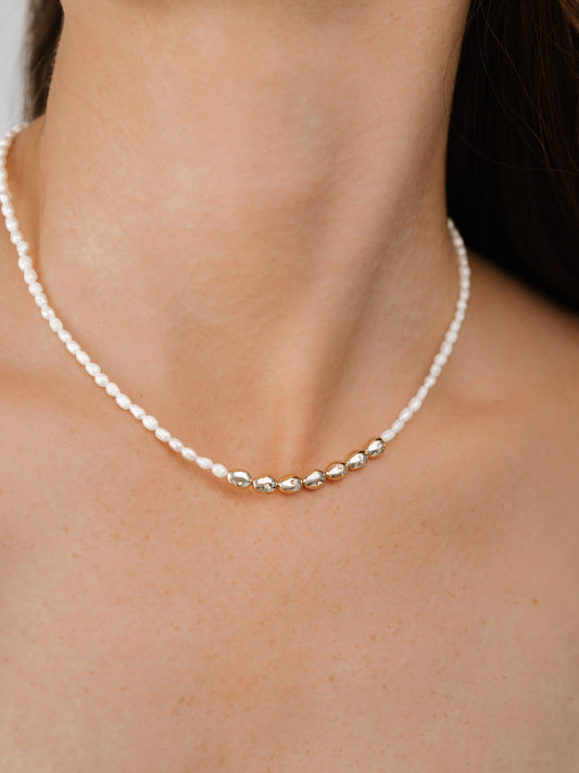 A delicate necklace rests on a person's neck, featuring alternating white beads and gold oval accents, against a background of smooth skin and dark hair.