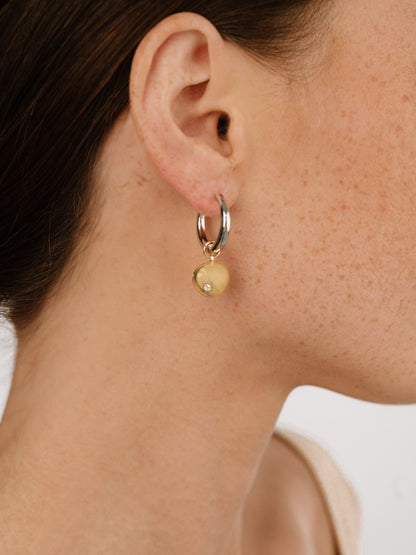 A close-up of an ear wearing a hoop earring with a large, round yellow pendant embellished with a small gem, set against a plain, light background.