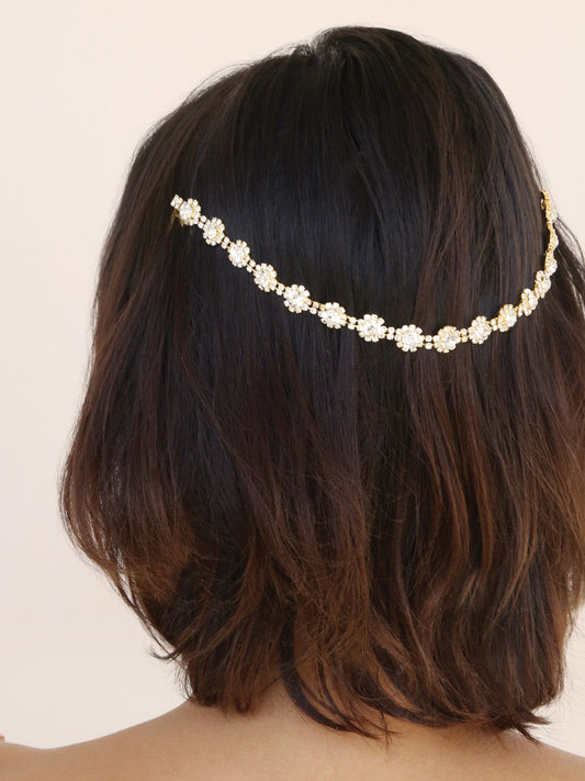 A gold and pearl hair accessory is positioned in wavy brown hair, enhancing its elegance. The backdrop is a soft, neutral tone, creating a calm and sophisticated atmosphere.