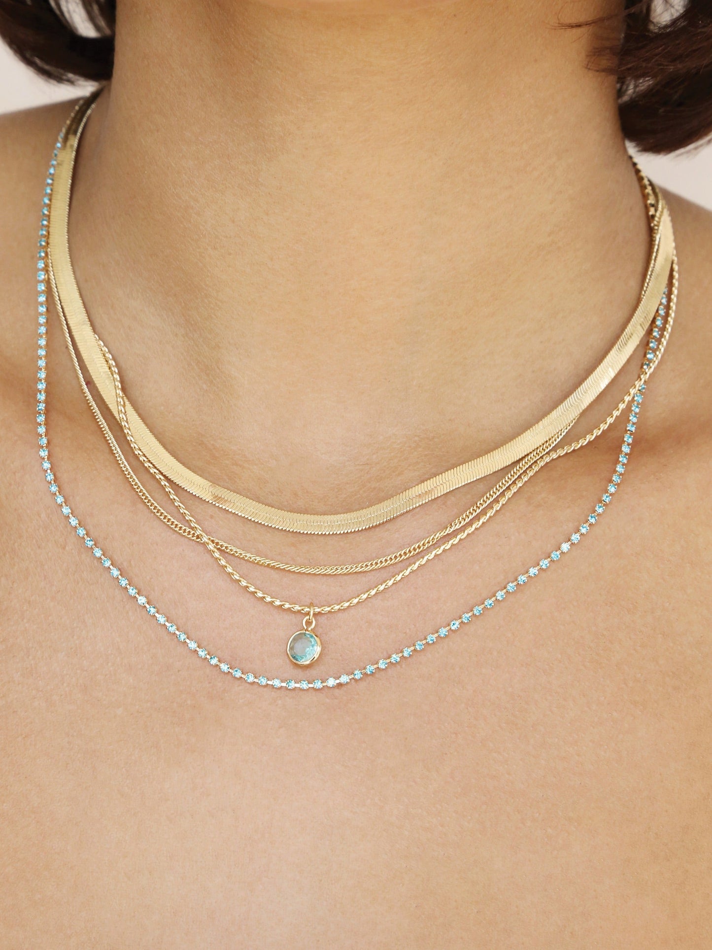 A layered collection of necklaces adorns the neck, featuring gold chains and a delicate blue gemstone. The jewelry sparkles against the skin in a well-lit setting.