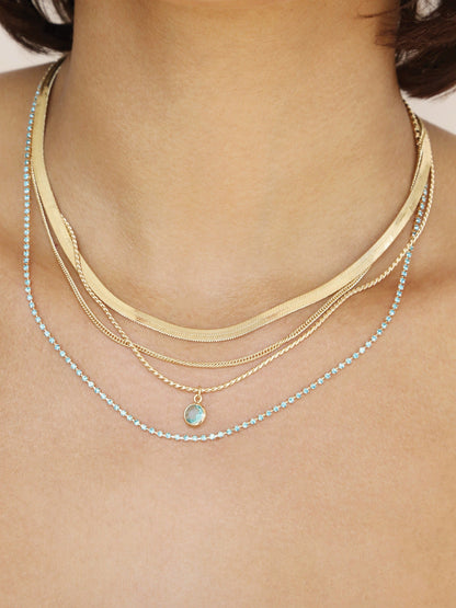 A layered collection of necklaces adorns the neck, featuring gold chains and a delicate blue gemstone. The jewelry sparkles against the skin in a well-lit setting.