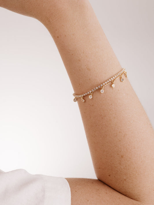 A delicate gold bracelet adorned with small charms hangs elegantly on a light-skinned arm, against a softly lit, neutral background that enhances the jewelry's sparkle.