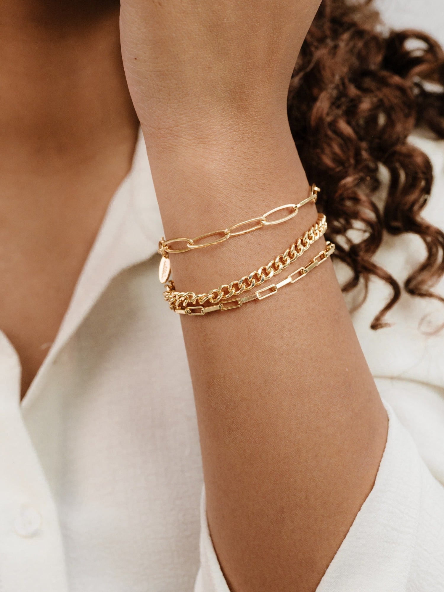 A golden bracelet set adorns a wrist, featuring different chain styles. The wrist is partially covered by a light-colored, textured shirt, with wavy hair blurred in the background.