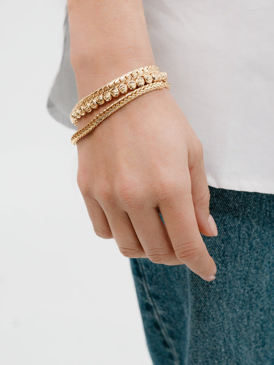 A hand is adorned with three gold bracelets of different designs, resting against denim fabric. The arm is positioned casually, complementing a light-colored shirt.