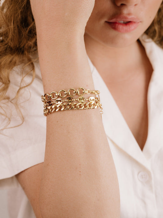 A womanâ€™s arm showcases multiple gold bracelets, with one hand raised to her face. She wears a white shirt, and her lips are slightly parted, suggesting a contemplative expression.