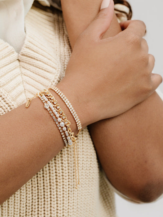 A wrist adorned with multiple elegant bracelets, including gold and pearl accents, is positioned against a textured, cream-colored sweater. The hand gently rests against the arm.