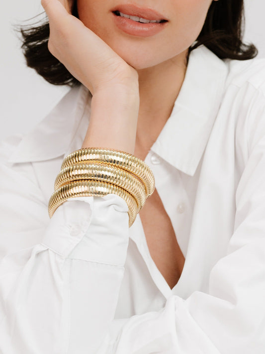 A woman's hand rests on her face, adorned with multiple gold-tone bangles, while she wears a classic white button-up shirt. The background is soft and neutral, enhancing the focus on her accessories.