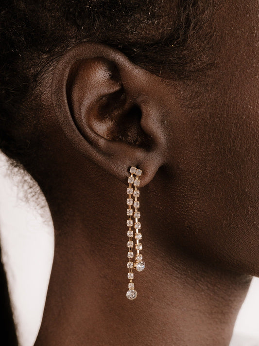 A long, elegant earring featuring sparkling crystals hangs from a pierced ear, accentuating the skin tone and texture of the model's neck and nearby hair.