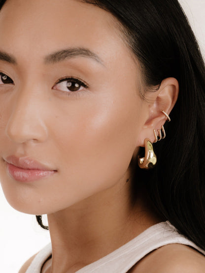 A young woman with long, dark hair is wearing gold earrings. She gazes slightly to the side, highlighting her makeup and the shiny jewelry against a softly lit, neutral background.