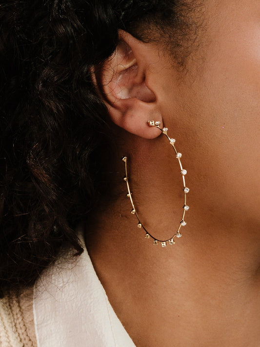 Gold hoop earrings adorned with small gems are worn on the ear of a person with curly hair, against a soft background that subtly blends with their skin tone.