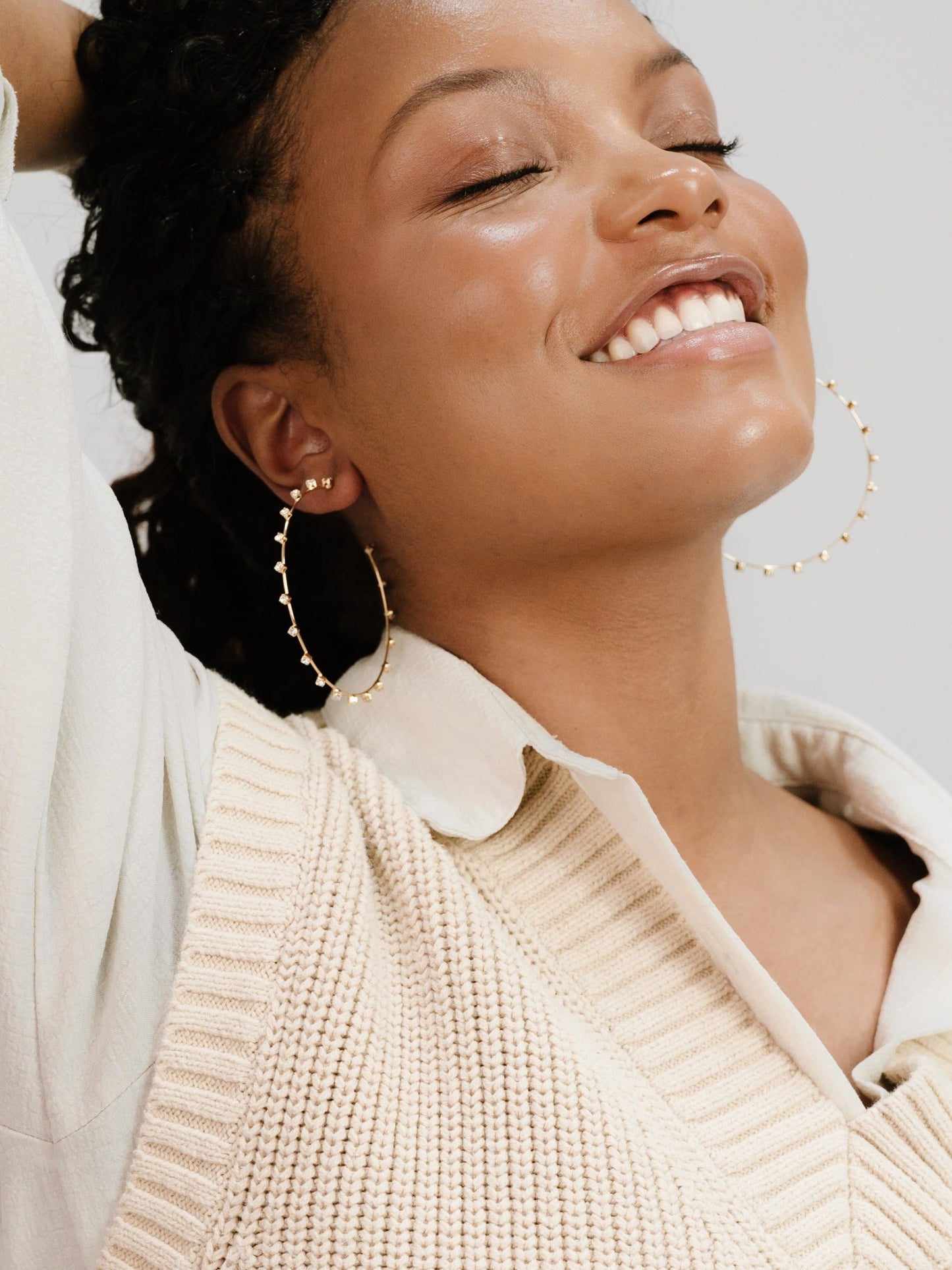 A woman with curly hair smiles joyfully, wearing large hoop earrings and a knitted beige sleeveless top over a collared shirt, relaxing against a soft, light background.