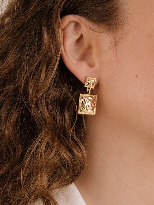 A gold earring hangs from a woman's ear, featuring a textured rectangular design. The woman has wavy, light brown hair and is in a softly lit, neutral-toned setting.